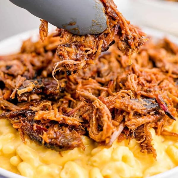 Tongs lifting saucy, shredded Phat Daddy's BBQ pulled pork from a bowl of creamy mac and cheese.