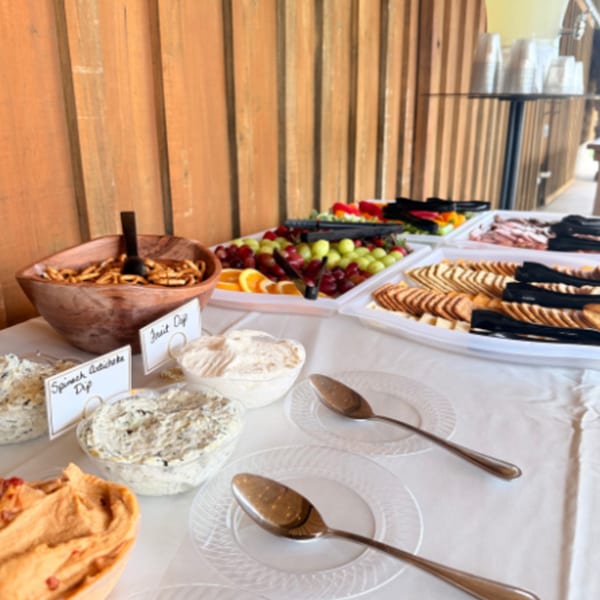 Buffet table with dips, crackers, pretzels, and fruit, set against a wooden wall.